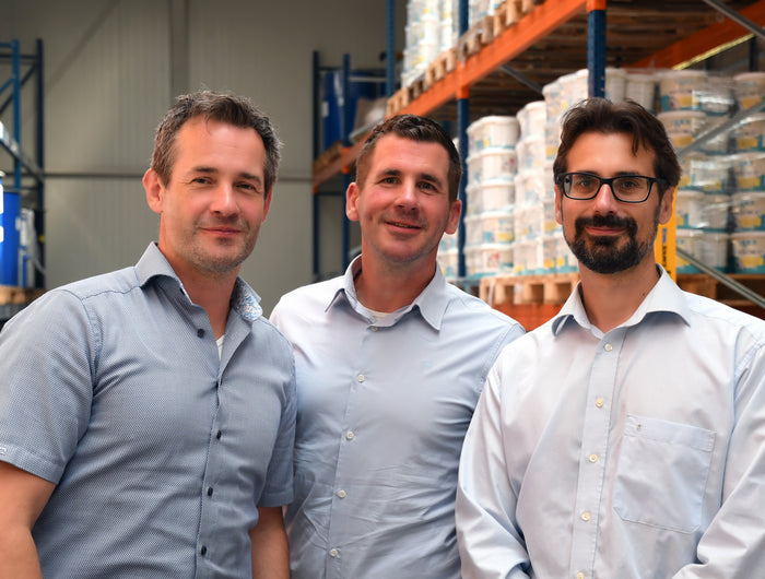 Three men standing in a warehouse with shelves and containers in the background